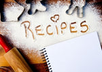 Butcher block counter with icing sugar dusted thickly over a large surface with the word "Recipes" drawing by hand and a heart shape dotting the "i" in "recipes". There are metal cookie cutters strewn around & a rolling pin & notebook to the side.